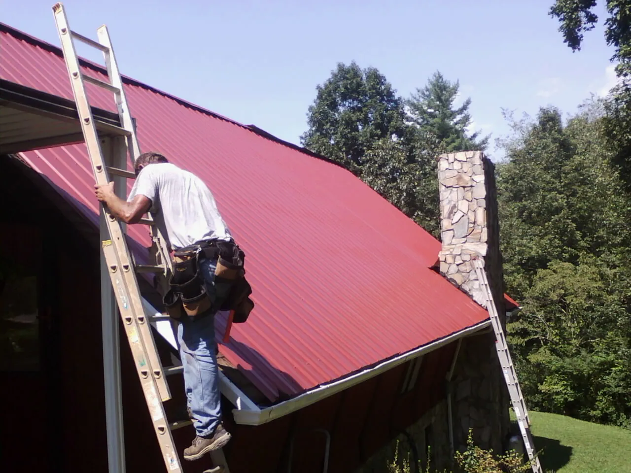 Roofer on a ladder beside a red metal roof and stone chimney — Soffit & Fascia Repair by Roofing Brothers in Toluca